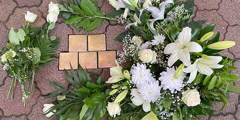 Blick von oben auf zweireihig im Boden verlegten goldenen Stolpersteine, um die Gestecke mit weißen Blumen und 3 einzelne weiße Rosen liegen.  