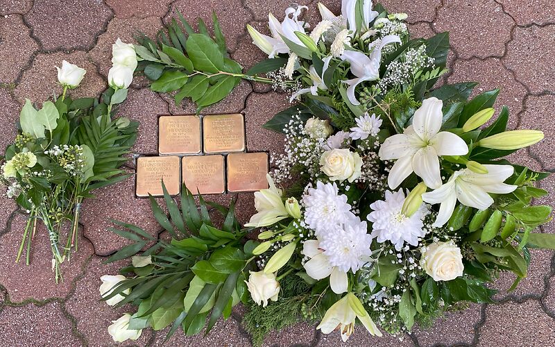 Blick von oben auf zweireihig im Boden verlegten goldenen Stolpersteine, um die Gestecke mit weißen Blumen und 3 einzelne weiße Rosen liegen.  