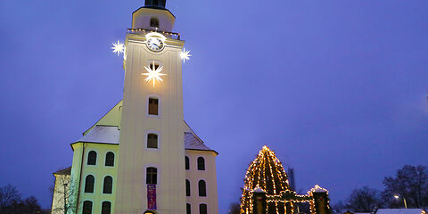 Links steht die beleuchtete Stadtkirche St. Nikolai mit 3 hellen Sternen am Glockenturm. Daneben ist der Eingang zum Forster Weihnachtsmarkt mit einem EIngangstor, mehreren Ständen und dem mit Lichterketten behangenden Weihachtsbaum zu sehen.