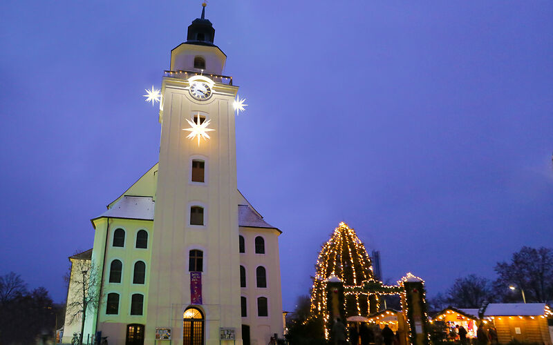 Links steht die beleuchtete Stadtkirche St. Nikolai mit 3 hellen Sternen am Glockenturm. Daneben ist der Eingang zum Forster Weihnachtsmarkt mit einem EIngangstor, mehreren Ständen und dem mit Lichterketten behangenden Weihachtsbaum zu sehen.