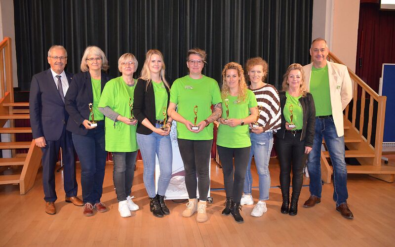 Vor der Bühne stehen mehrere Personen in einer Reihe, die meisten von ihnen tragen grüne Shirts. Bis auf die äußeren Personen hält jeder eine goldene Auszeichnung in der Hand.