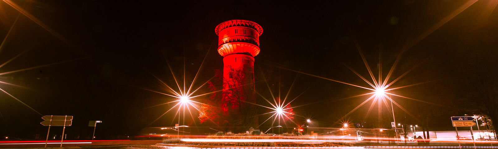 Mittig im Bild ist der rot angestrahlte Wasserturm bei Nacht zu sehen. Um ihn herum leuchten 3 Laternen.