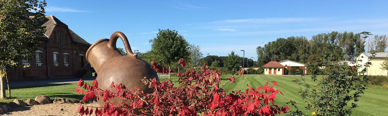 Hof im Gut Neu Sacro, blauer Himmel, ein übergroßer schrägstehender Krug aus gebranntem Ton, herbstlich dunkelrotem Laub eines Strauches