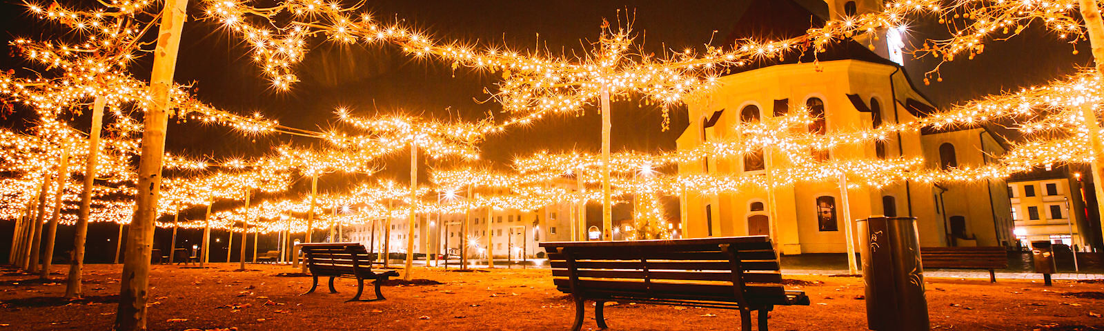 Zwischen Bäumen sind im Schachbrettmuster leuchten Lichterketten gespannt, darunter stehen zwei Bänke. Im Hintergrund befindet sich die Stadtkirche.