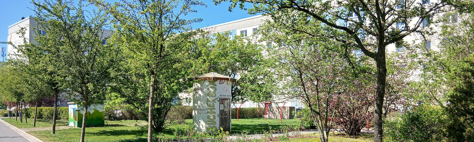 Die Bücherbox umgeben von blühenden Bäumen vor einem mehrstöckigen Wohnhaus.