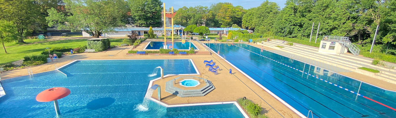 Blick aus einer erhöten Position auf das Schwimmerbecken (rechts) und das Nichtschwimmerbecken (links), dahinter der Sprungturm mit Becken. Hinter den Becken ist Wiese.