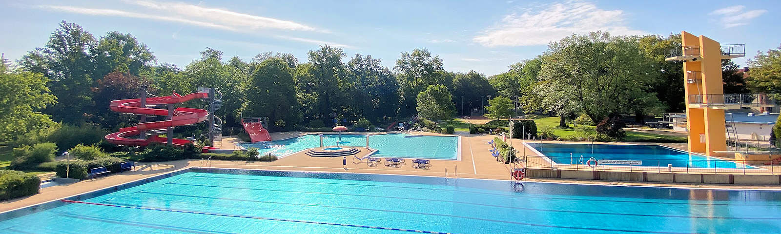 Vorn befindet sich das Schwimmerbecken, dahinter befinden sich rechts die Rutschen mit dem Nichtschwimmwerbecken und links der Sprungturm mit Becken. Zwischen den Becken führ ein gepflasteter Weg entlang. Im Hintergrund stehen viele große Bäume. Die Sonne scheint.