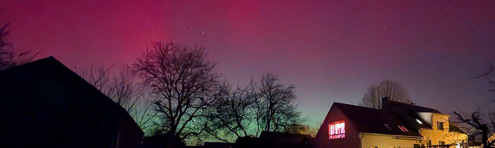 Der Himmel leuchtet in einem Farbverlauf von pink zu grün, im Vordergrund sieht man die Silhouetten von Bäumen und ein beleuchtetes Haus.