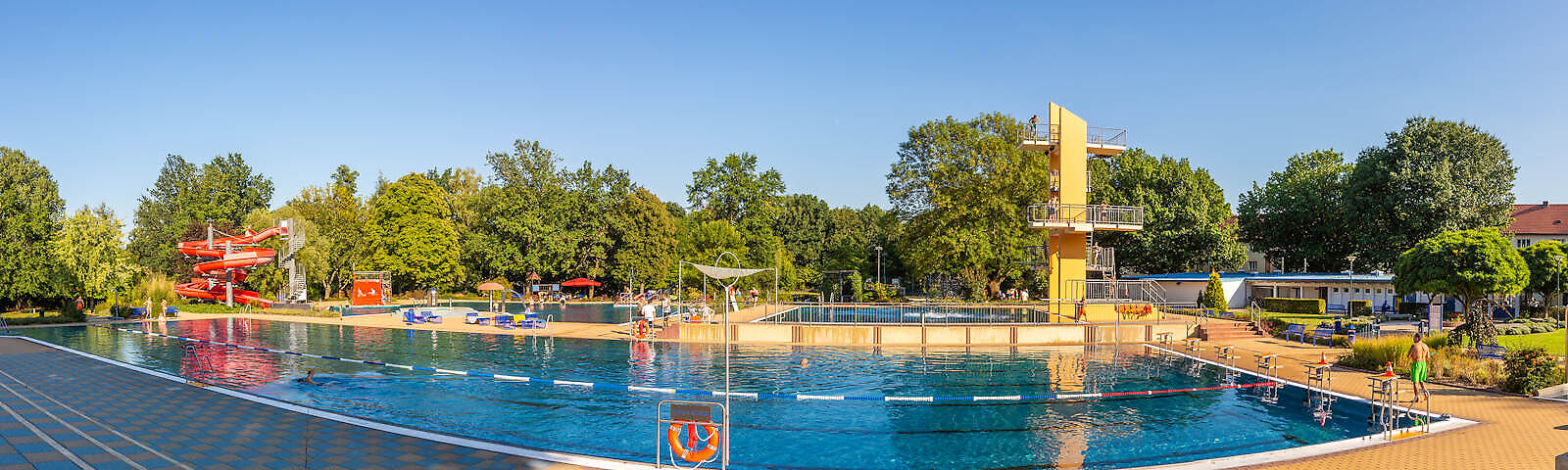 Zu sehen ist das Freibad mit seinen Wasserbecken und die Grünanlagen, die Liegewiesen und der Sprungturm.