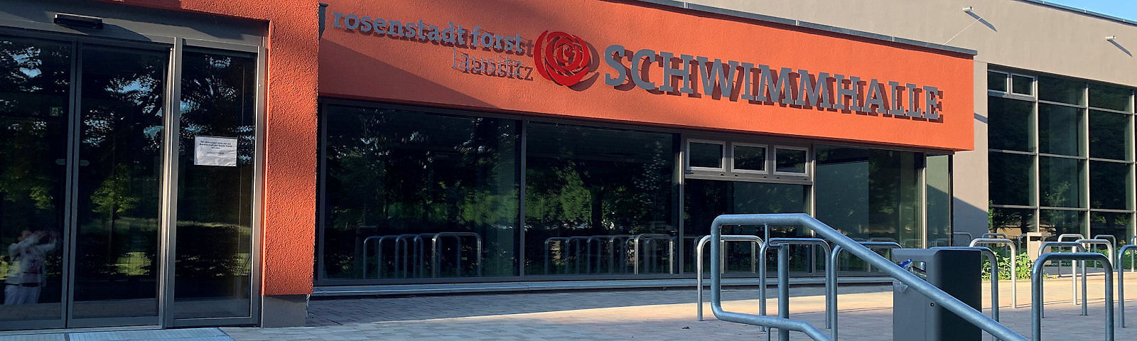 Blick auf die Schwimmhalle von außen: Links ist die gläsernde EIngangstür, die Wand rechts davon besteht im unteren Teil aus Fenstern, darüber ist sie rot gestrichen und in grauen Buchstaben steht rosenstadt lausitz SCHWIMMHALLE dran, dazwischen ist eine Nachbildung einer roten Rose 