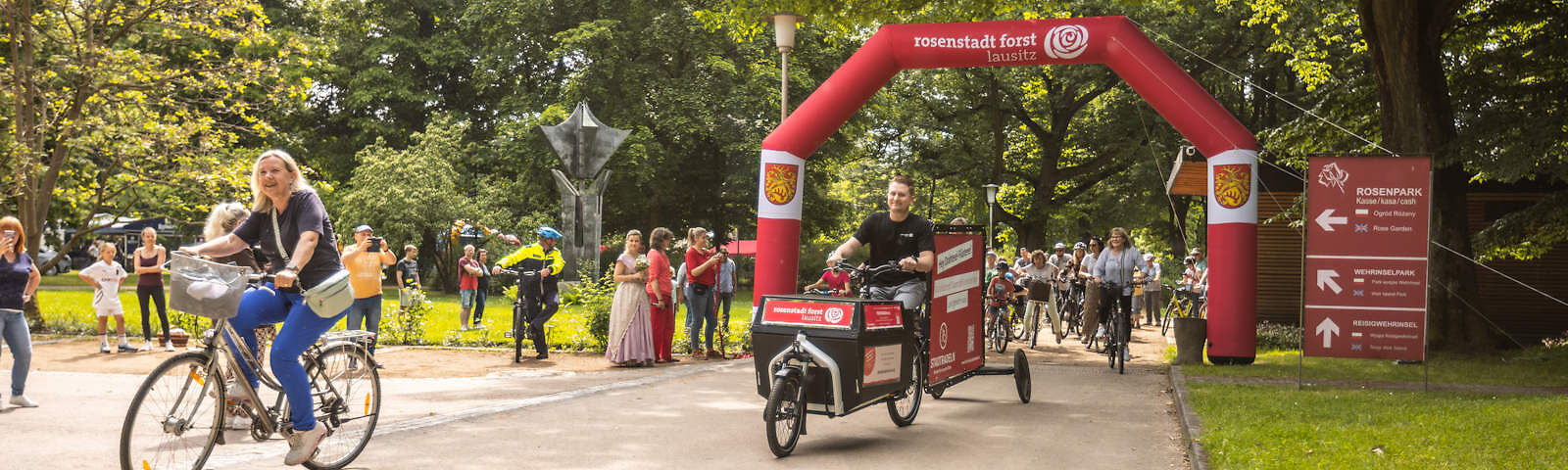 Auf einem Sandweg im Rosengarten wird das Stadtradeln eröffnet. Bürgermeisterin Simone Taubenek fährt voran, schräg hinter ihr ist ein umgebautes Rad mit Werbetafeln und Werbebannern, weitere Teilnehmende sind hinter einem roten Bogen mit der Aufschrift rosenstadt forst lausitz zu sehen.