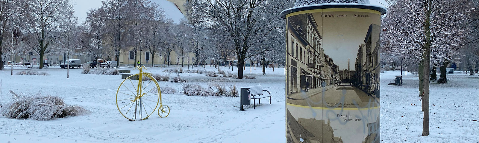 Der verschneite Park hinter der Stadtkirche mit einer Säule auf der historische Bilder von Forst gezeigt werden, einem gelben historischen Fahrrad und der Stadtkirche im Hintergrund.