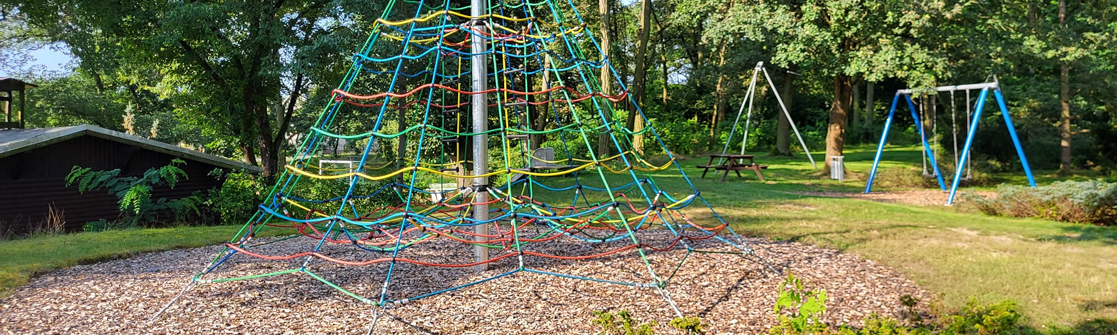 Ein Kletterturm mit bunten Seilen und eine Duppelschaukel, ein Tisch mit Bänken im Hintergrund